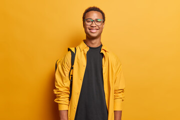 Wall Mural - Portrait of delighted cheerful guy with short dark hair dressed in casual clothing poses with backpack has travel lifestyle poses against bright yellow wall feels slef proud after success on session