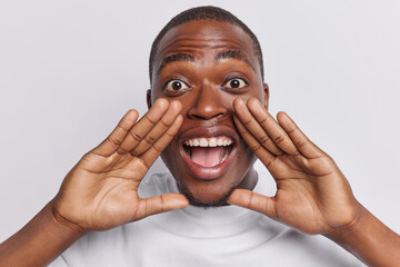 Wall Mural - Close up shot of emotional dark skinned man exclaims loudly keeps hands near widely opened mouth calls someone dressed in casual clothing isolated over white background. People and emotions.