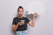 Horizontal shot of shocked Latin woman browses her smartphone holds blank communication bubble for your advertising content wears casual black t shirt and jeans isolated over white background
