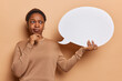 Thoughtful young African woman holds chin dressed in casual jumper holds speech bubble for your advertising content dressed in casual jumper isolated over brown background. Place your ideas here