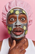 Headshot of dark skinned man has bugged eyes and opened mouth applies nourishing clay mask with cucumber slices undergoes beauty procedures at home wears bath hat isolated over pink background