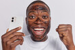 Positive dark skinned man clenches fist and smiles gladfully celebrates good news holds mobile phone texts online applies facial scrub mask for skin treatment isolated over white background.