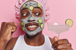 Close up shot of dark skinned man looks at cocktail clenches fist has surprised cheerful expression applies beauty mask and cucmber slices undergoes beauty procedures isolated over pink background