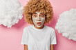 Photo of scared fair haired woman stands speechless applies facial clay mask for reducing fine lines has bugged eyes feels shocked wears casual white t shirt isolated over pink background white clouds