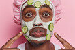 Close up shot of shocked dark skinned adult man with beard applying cucumber mask on his face points index fingers at cheeks wears bath hat isolated over pink background. Beauty and spa concept.