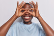 Funny silly playful carefree dark skinned guy having fun fool around mimicking show goggles looks through finger made glasses smiles toothily dressed in casual clothes isolated over white background.