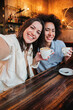 Vertical portrait of two young adult women having fun taking a selfie at coffee shop sitting at table with cups. Pair of joyful females smiling at restaurant. A couple of girls enjoying together. High