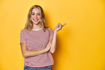 Wall Mural - Young blonde Caucasian woman in a red striped t-shirt on a yellow background, smiling cheerfully pointing with forefinger away.