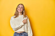 © Asier - Young blonde Caucasian woman in a white sweatshirt on a yellow studio background, smiling and pointing aside, showing something at blank space.