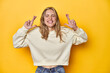 Young blonde Caucasian woman in a white sweatshirt on a yellow studio background, crossing fingers for having luck