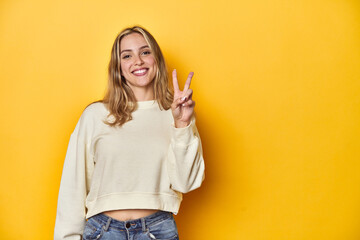 Young blonde Caucasian woman in a white sweatshirt on a yellow studio background, showing victory sign and smiling broadly.