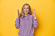 Young blonde Caucasian woman in a violet sweatshirt on a yellow background, celebrating a victory or success, he is surprised and shocked.