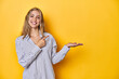 © Asier - Young blonde Caucasian woman in a striped business shirt on a yellow background, excited holding a copy space on palm.