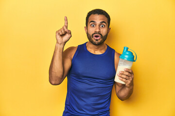 Wall Mural - Athletic young Latino man holding a protein shake against a yellow background having some great idea, concept of creativity.