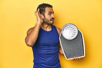 Wall Mural - Fit young Latino man with scale on yellow background, fitness concept, trying to listening a gossip.