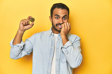 Wall Mural - Young Latino man holding a Bitcoin coin, yellow studio background, biting fingernails, nervous and very anxious.