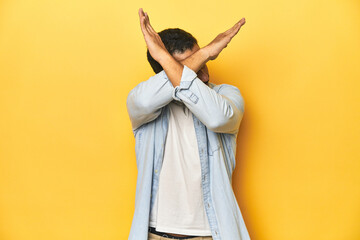 Wall Mural - Casual young Latino man against a vibrant yellow studio background, keeping two arms crossed, denial concept.