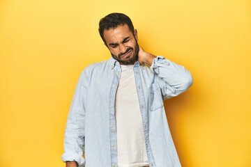 Wall Mural - Casual young Latino man against a vibrant yellow studio background, having a neck pain due to stress, massaging and touching it with hand.