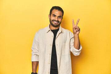 Wall Mural - Casual young Latino man against a vibrant yellow studio background, showing victory sign and smiling broadly.