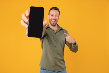 Excited caucasian man holding blank cellphone showing screen to camera pointing at it, recommending website or application, mockup banner