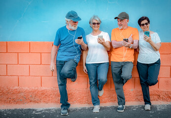 Cheerful group of senior people leaning against an orange and blue wall wearing earphones looking at their phones enjoying technology and social