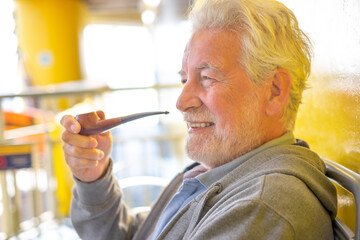 Sticker - Side view portrait of smiling relaxed senior white haired man sitting in outdoor smoking pipe. Elderly bearded grandfather enjoying free time and smoke