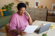 © Seventyfour - Smiling aged businesswoman with financial document talking to her employer on mobile phone while sitting by desk in front of laptop