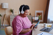© Seventyfour - Side view of smiling mature woman in headphones and pink t-shirt looking through playlist in smartphone while choosing something to listen