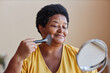 © Seventyfour - Smiling African American woman touching her face with brush while applying loose powder on skin and looking in mirror during makeup
