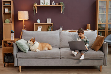 Wall Mural - Wide angle view of little boy with down syndrome using laptop in cozy home interior sitting on soft couch with cute dog, copy space