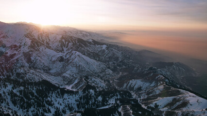 Wall Mural - Epic red-orange sunset in high snowy mountains. There are tall coniferous trees, there is snow on the hills. High peaks. The sun's rays fall on the clouds and fog that is lower down the gorge. Almaty