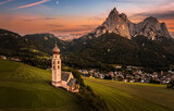 Fototapeta Londyn - Seis am Schlern, Italy - Aerial panoramic view of St. Valentin Church and famous Mount Sciliar mountain at background with colorful sunset sky and warm sunlight at South Tyrol on a summer afternoon