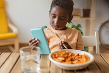 Boy Eating Food And Using Smart Phone At Dining Table