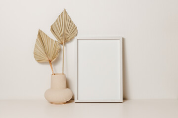 Wall Mural - Empty aesthetic photo frame mockup with vase and palm leaves on white table. Picture frame template with copy space for poster and design, home interior decoration, cozy scandinavian style