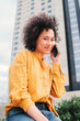 © Jose Calsina - Vertical portrait of young latin woman with curly hair having a call conversation with a smartphone. Hispanic happy lady with yellow shirt talking by cellphone sitting outdoors. Female using phone