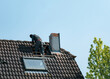 A roofer stands atop a house, destroying the chimney from an unusually low angle view. Its distinctive architecture and tiled roof stand out against the clear sky.