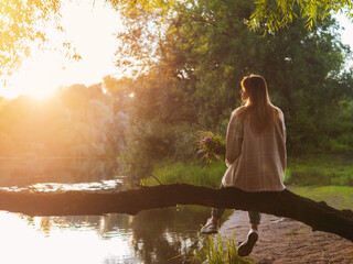 A young woman sits on a tree trunk above the river with a bouquet of wildflowers and enjoys the sunset. Summer time