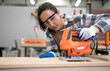 © Nassorn - Beautiful Latin woman carpenter using power tools working her wood job in carpenter's shop. Young hispanic female in protective goggles busy in furniture woodworking. Feminism in carpentry industry.