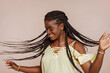 © carlesmiro - Young happy smiling african woman celebrates and enjoys herself with a hair flip, wears braids and yellow top, dancing at studio over beige background.
