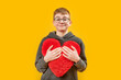 Smiling brunette boy in glasses holds plush red heart. Portrait of teenager with pillow in shape of heart on yellow background.