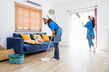 Two Asian Young Professional Cleaning Service Women Worker Team Working In The House. The Girl Housekeeper Wiped A Wet Mop On Wooden Floor With Another One Clean The Curtain With A Feather Duster.