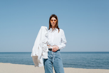 Stylish caucasian young woman in sunglasses posing on the beach on a sunny day. A slender woman standing on the shore looking at the camera