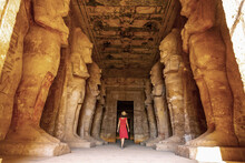 A Young Woman In A Red Dress At The Abu Simbel Temple Next To The Sculptures, In Southern Egypt In Nubia Next To Lake Nasser. Temple Of Pharaoh Ramses II, Travel Lifestyle