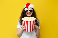 Young Girl In Santa Hat And 3d Glasses Holds Popcorn And Smiles On Yellow Isolated Background, Woman Viewer
