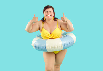 Wall Mural - Portrait of happy smiling fat woman in beach rubber ring wearing yellow swimsuit showing thumb up sign isolated on a blue studio background. Travel, vacation and summer journey concept.