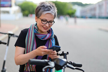 Poster - Smiling mature woman using smartphone to rent an electric scooter in the city.