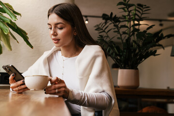 Wall Mural - Woman drinking coffee and using mobile phone while sitting in cafe