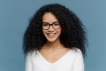 Curly woman with smile, glasses, white jumper, isolated on blue background. Expresses good emotions, enjoys day with friends.