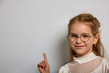 Pretty Schoolgirl In A White Blouse And Round Glasses Points Her Finger At The School Blackboard. Little Girl In Round Glasses With A Black Frame Stands Against A White Wall, Advertising Something