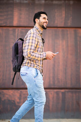 Wall Mural - Cheerful young arabic guy walking outside with bag and mobile phone